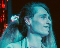 Heidi, a white nonbinary person with long brown hair tied up in an LED scrunchie, is smiling on stage in blue lights.