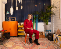 A person with blonde hair wearing a red boilersuit sitting on an orange stall in the middle of a white walled room with a navy corrugated metal panel behind them and pink patterned carpet under their feet. They are surrounded by an orange cupboard, big plant, various tools including a sewing machine and conductive objects they have made hang from the ceiling and sit at the sides of the room 