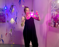 Suzi Bratt, white female, early thirties with pink hair and an arm of floral tattoos stands amidst a hanging sculpture of hands and metal arcade grabbers.