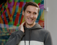 A white man with short, brown hair, wearing a green and white hooded jumper. He is standing up in front of an audience and smiling widely at an audience member