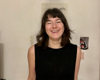 A white woman in her 20s with shoulder length brown hair, wearing a black sleeveless top.