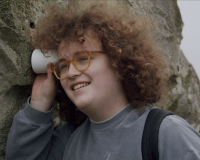 Robin Thorn, a young white person with long curly brown hair is wearing yellow glasses and has their head pressed against a small white cylindrical device that is touching an avebury stone.
