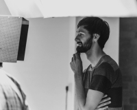 Reuben Armstrong, a white man with short dark hair and a beard, smiling looking at a Director's monitor whilst working on set.