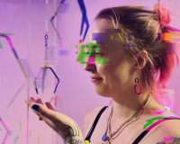 A glitched photo of Suzi Bratt, white female, mid thirties with pink hair and an arm of floral tattoos stands amidst a hanging sculpture of hands and metal arcade grabbers.