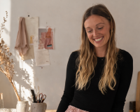 A image of Nina Redman in her Fashion Studio in Bristol, natural light coming into the windows and designs of clothing and samples of fabric on the table in front of her.