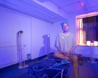 a photo of a person in a striped t-shirt and shorts sitting on a blue bench in the centre of a dark room. They are surrounded by wooden statues and the photo is double-exposed giving them a ghostly appearance.