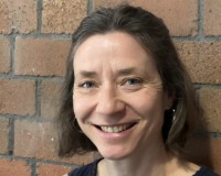 White woman with shortish brown hair and blue eyes, stood in front of a red brick wall