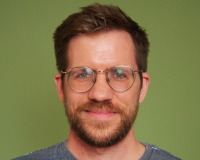 A white man in his mid thirties, with a beard and glasses. He is smiling and standing in front of a green background.