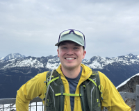 Smiling person wearing a bright yellow jacket and running vest standing in front of a view over a snowy mountain range.
