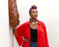 A Somali Woman in a red shirt and blue, yellow and red headwrap next to a grass weaving looking to the distance. 