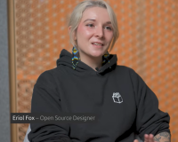An image of Eriol who is a femme presenting nonbinary person. They are a white person with tattoos on their left arm and wear a black Hoodie with a Penpot logo, have blonde hair and wear Kenyan beaded earrings that are black, blue and yellow