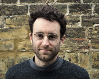 A man with dark hair and silver glasses stands in front of a brick wall. the man is wearing a woollen jumper and is smiling.