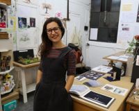 Photograph of Alison standing in her studio, surrounded by sketches and theatre model boxes.