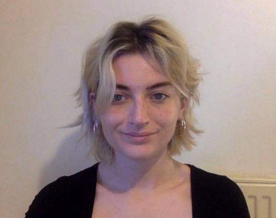 Woman with short curly blonde hair and black top against beige wall