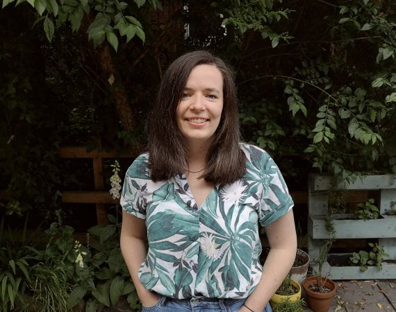 Corinne is standing in a garden with dark brown mid length hair and tropical shirt.