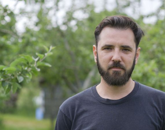 Lukus is a white male with a dark beard and hair. He is wearing a black t-shirt and is standing in front of a number of trees in summer time.