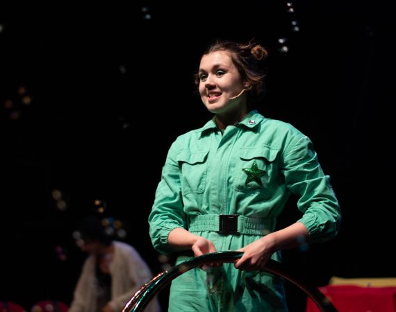 Scarlett stands just right of the centre holding two hula hoops by her waist. She is smiling, surrounded by bubbles and wearing a green boiler suit with a sequin star on the chest pocket. 