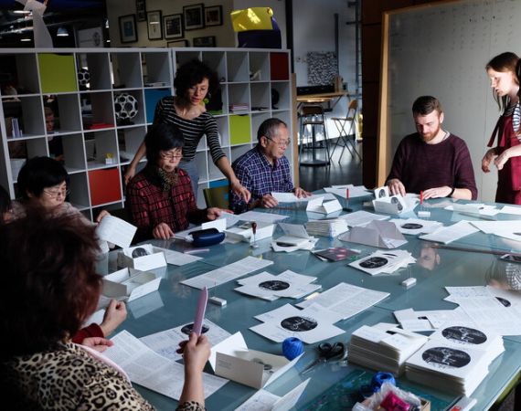 A group of men and women, young and old, seated and standing around a table. They are hand-finishing the Here We Grow books.