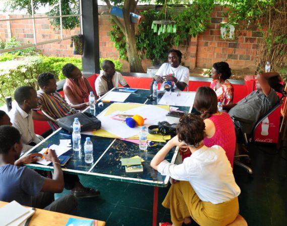 A group of black men and women seated around a table. In the centre of the table are items to make books - paper, string. There are also headphones on the table. Surrounding the people are plants and trees.  