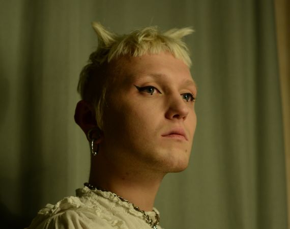 Marcin is a white, androgynous person with short blond hair. He is wearing black eyeliner, a ruffled dress and a chain with a padlock on his neck.