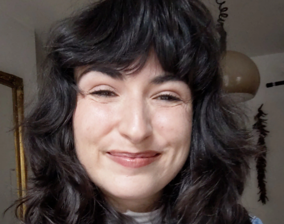 A 30-year-old woman with dark brown hair smiling and squinting, wearing a green shirt under a blue denim shirt