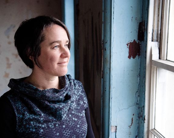 White female with short dark hair looking out of window smiling wearing a patterned blue dress with a large roll neck