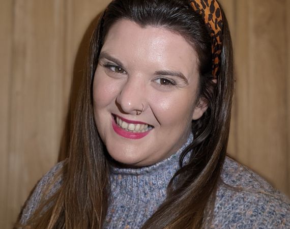 An image of Liz's head and shoulders. She is a white woman and is smiling. She has long brown straight hair and is wearing a leopard print headband and a fuzzy purple jumper.