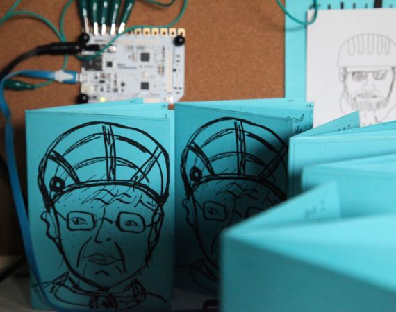 A blue booklet with a drawing of a person wearing a helmet is wired up to a conductor to make sound when touched.