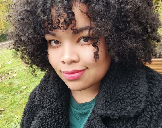 Malaika is a mixed race (Black African & White) cis woman. She has shoulder length dark, curly, afro hair with a fringe. She is wearing a green jumper dress with a fluffy black coat over the top. She smiles at the camera, with light red lipstick on.