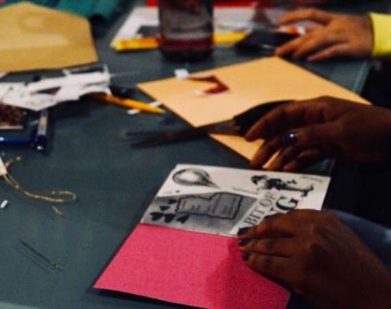 A black woman, seen only by the hands, customises and designs her zine which lays open on a dark green table surrounded by scissors, colourful prints, stamps, pins and other crafting material.