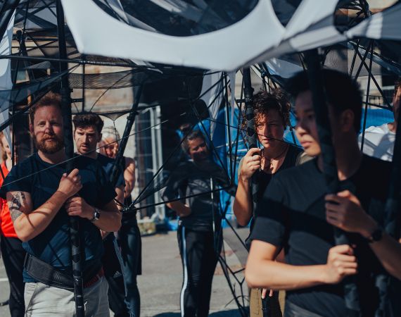 6 people holding up an abstract structure. They are at different distances from the camera