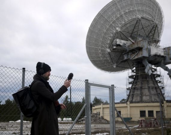 An image of Duncan Speakman dressed all in black, with a black hat, holding a smartphone taking a picture of a large grey satellite