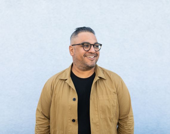 An Image of Nikesh Shukla wearing a yellow shirt with a black t-shirt underneath, he is looking into the distance and smiling.