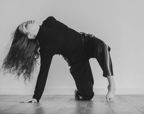 A dancer arches their back with both feet and one hand on the floor. The picture is in black and white. The dancer is wearing dark clothing and has long dark hair. The floor is shiny and wooden