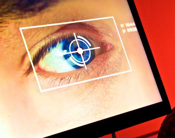 An image of a TV screen, in front of a red wall. On the screen is a close up of someones eye, over the top of the eye is a a graphic that appears to be scanning the blue iris of the eye