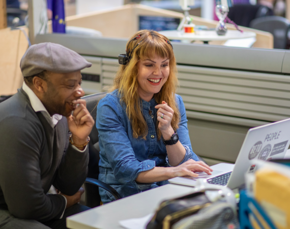 Two people sit on chairs at one of the hot desks at Pervasive Media Studio. They both look towards a laptop that sits on the desk smiling.