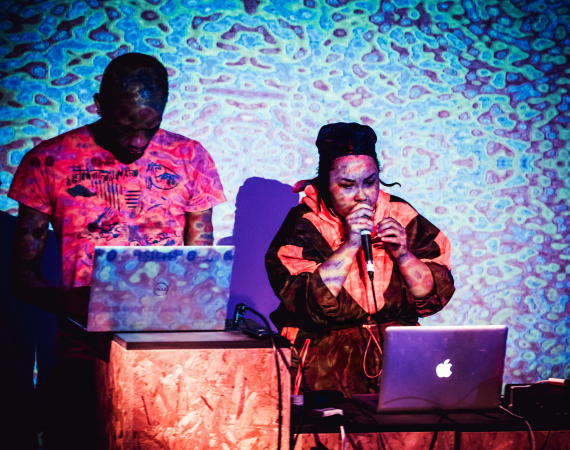 an image of two people looking at their laptops, one is speaking into a microphone, psychedelic visuals are project over them and the wall behind