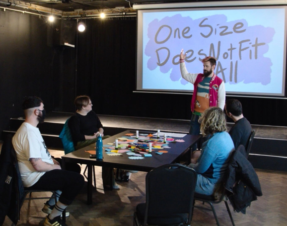 Still from a film of Graham speaking to 4 people sat at a table. A presentation says One Size Does Not Fit All in the background