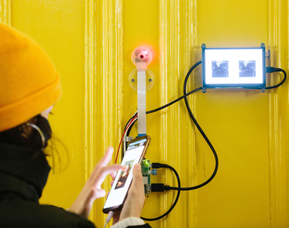 Photo of someone wearing a yellow hat, taken from behind, in front of a yellow door holding a mobile phone connected to another screen on the door