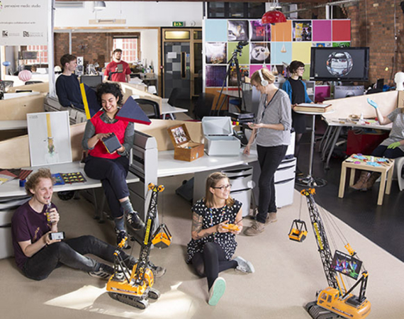 A photo of residents posing in the Pervasive Media Studio next to various projects including robotic cranes