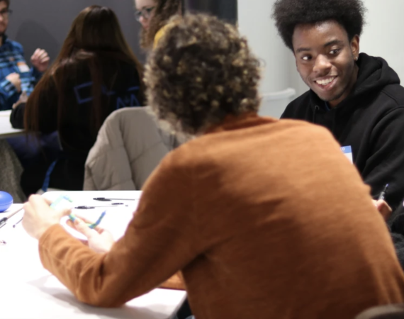 Two people are in the forefront of the image, sat around a table having a conversation. Another two people are in the background of the image, also sat around a table having a discussion.