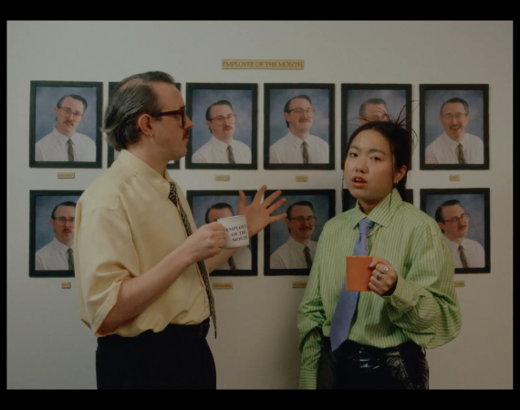 a still from a music video. Two colleagues appear to be stood in front of an employee of the month picture frame wall. The pictures are all of the same man