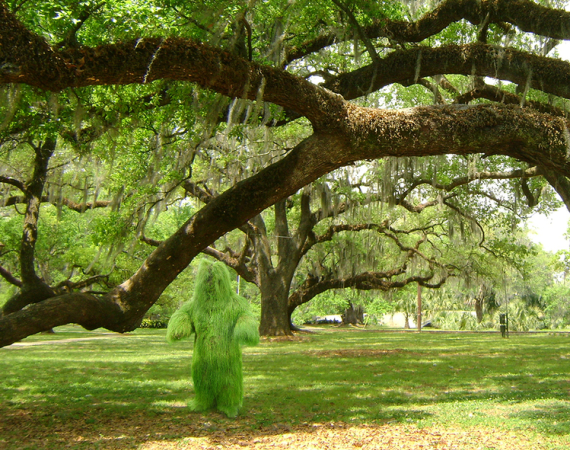 A photo of a person covered in a living grass costume made by Ashley Peer stood under a tree on grass