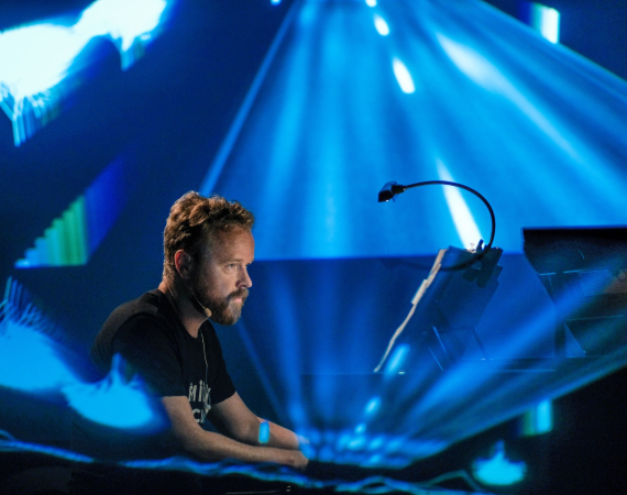 Pianist and composer Larkhall performs on piano, awash with blue lighting within a dark venue.