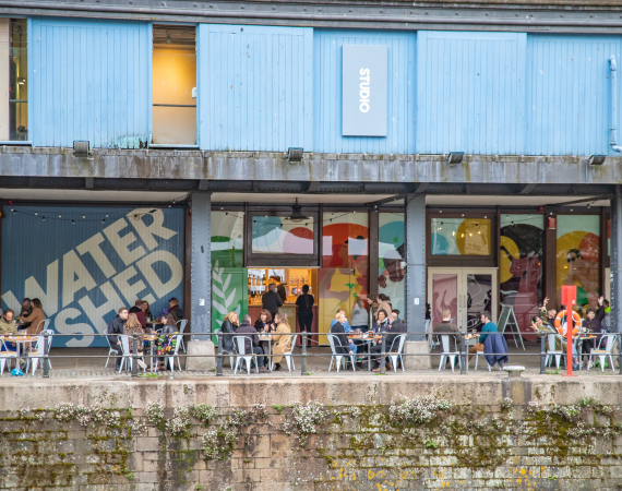 A photo of the external area of Watershed, focusing on the Undershed. Large illustrated artwork by Lucy Turner is on the windows and people sit around tables outside on the Harbourside