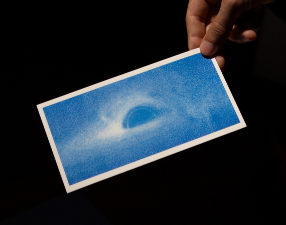A hand holding a baby blue postcard depicting a black hole, against a black background.