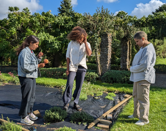 Three people stand outdoors looking at the ground, grass covered by tarpaulin