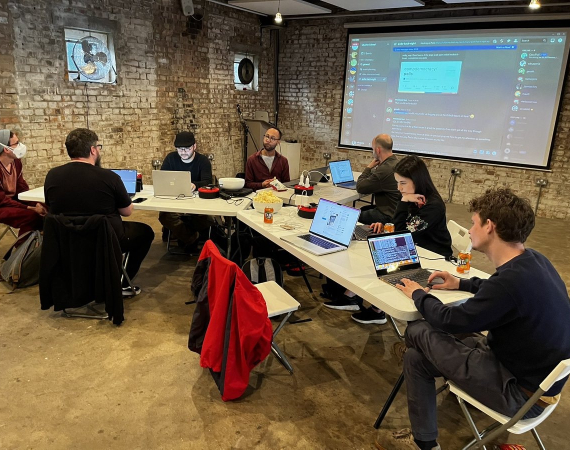 An image of of various people at a shared desk in a triangle formation, working on creative projects collaboratively. 