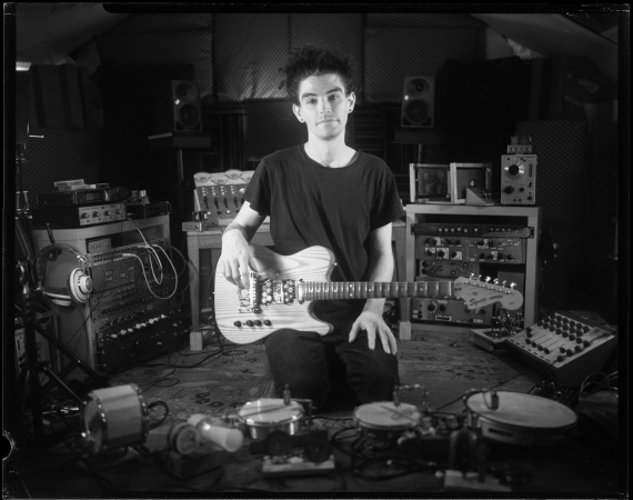 An image of instrument builder Craig Scott with his home built guitar, in black and white. He is surrounded by various different types of musical paraphernalia. 