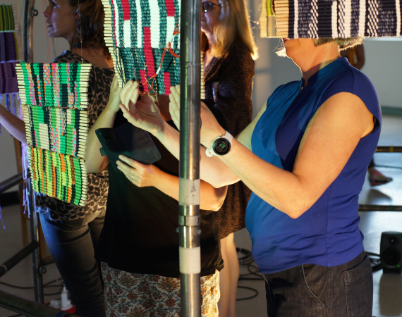 3 people explore woven tapestries suspended from a metal frame